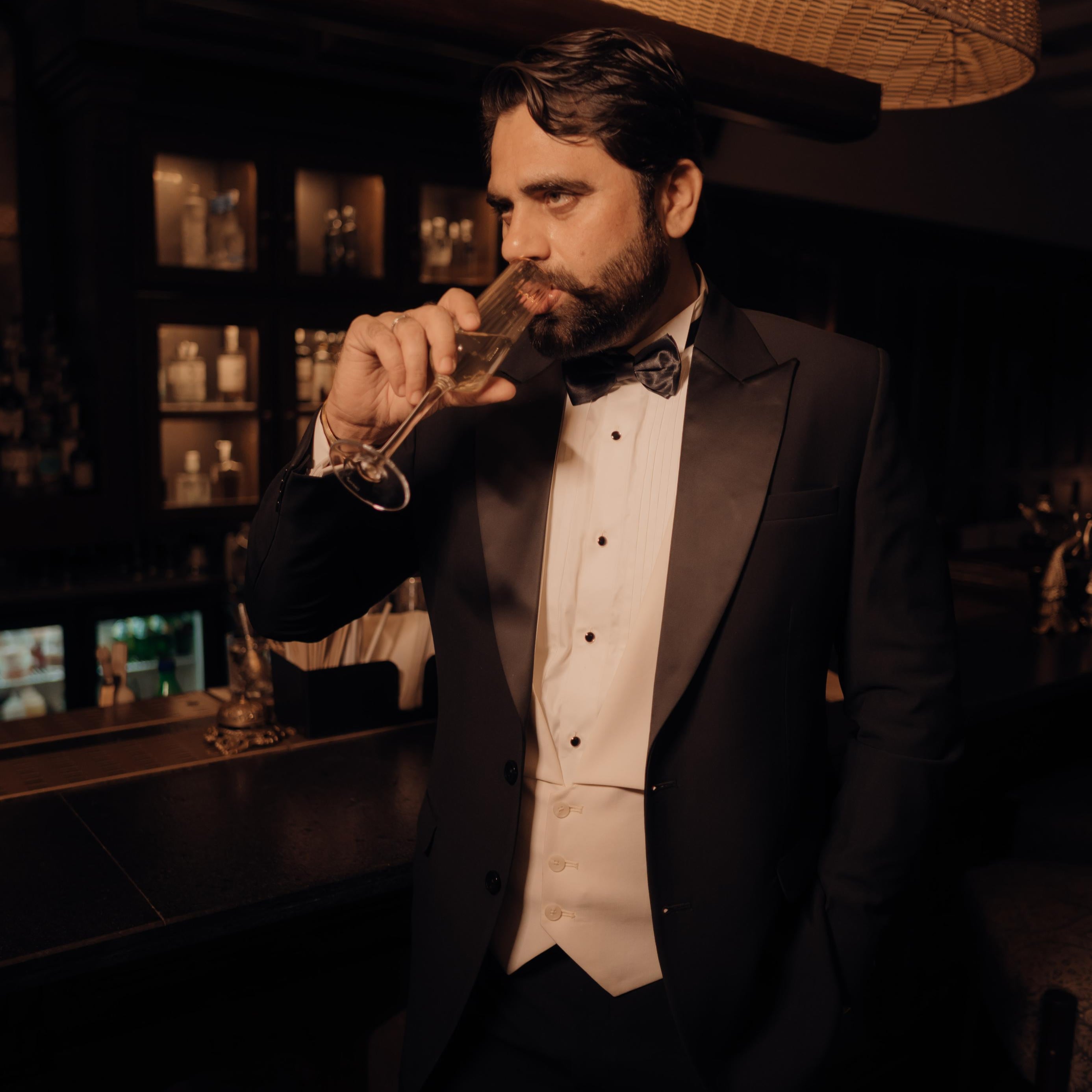 Man in a tuxedo drinking from a glass in a bar setting